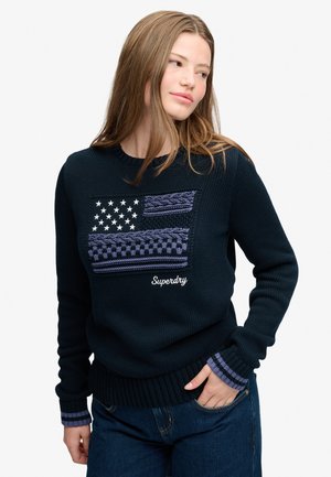 Ung kvinde med langt hår bærer en mørkeblå trøje med en tekstureret amerikansk flag-design og "Superdry"-logo, kombineret med blå jeans.