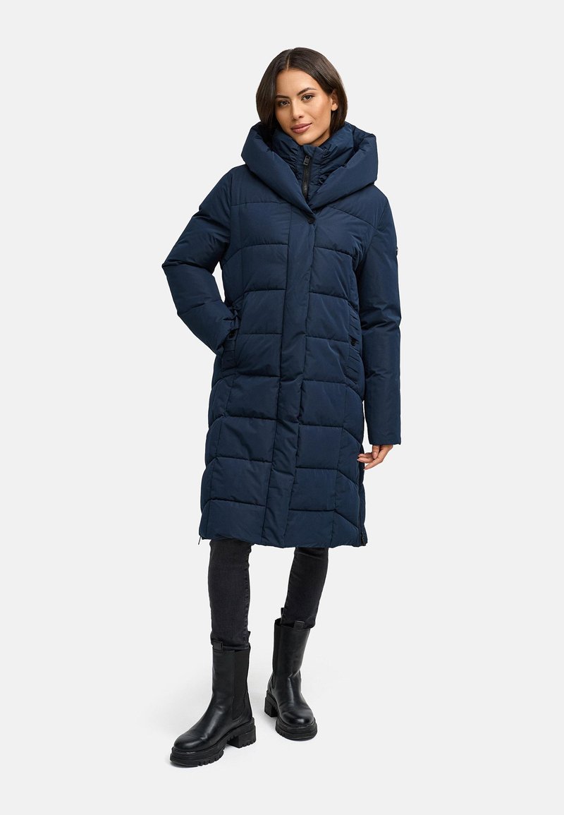 Marineblauwe, lange gewatteerde jas met een grote capuchon, horizontaal gestikte design en zijzakken, gecombineerd met zwarte enkellaarsjes en donkere broeken.