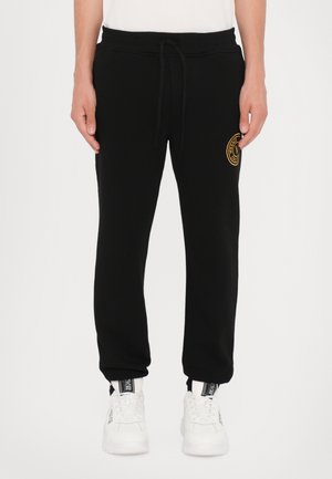 Black sweatpants with a drawstring waistband, featuring a gold circular logo on the left thigh and tapered cuffs at the ankles.