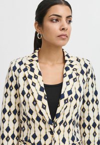 Bílý blazer s modrým a žlutým diamantovým vzorem, se zářezovým límcem, jednobotonovým zapínáním a hladkým povrchem. Černý top pod ním.