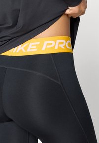 Leggings negre cu o bandă galbenă largă pe care scrie "NIKE PRO" în text alb. Fabricate dintr-un material neted și elastic, cu cusături detaliate.