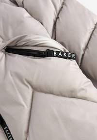 Giacca puffer beige chiaro con un design a motivi diagonali, zip frontale con un tirante nero "BAKER" e una texture liscia e satinata.
