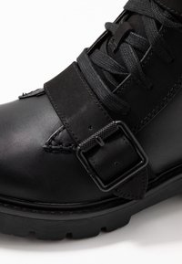 Czarny skórzany but z teksturowaną cholewką, metalowym paskiem z klamrą, czarnymi sznurowadłami i solidną gumową podeszwą, podkreślający trwałość i wsparcie.