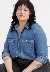 Camicia in denim blu con bottoni, dotata di due tasche frontali, maniche arrotolate e una consistenza morbida. Caratterizzata da un colletto classico e dettagli in bianco sui bottoni.