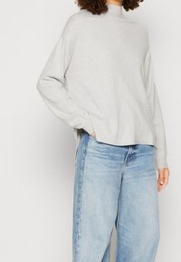 Ljusgrå, långärmad tröja med mock-neck, avslappnad passform och sidospalter. Bärs med högmidjade ljusblå jeans. Mjuk textur.