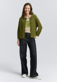 Olivgrüne Rippstrickjacke mit Rundhalsausschnitt und vorderen Knöpfen, kombiniert mit einem hellbeigen Grafik-T-Shirt und dunklen, ausgestellten Jeans.