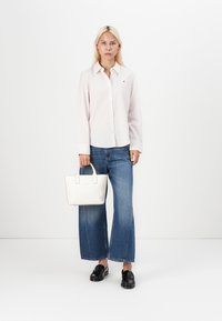 Camicia button-up a maniche lunghe rosa chiaro, jeans blue a gamba larga, mocassini neri, che tiene una piccola borsa bianca. Design e palette di colori minimalisti.