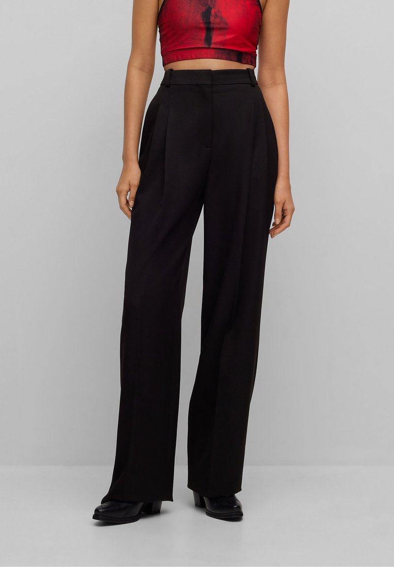 Femme portant un pantalon noir taille haute à jambes larges, des chaussures noires et un haut court sans manches rouge sur un fond gris uni.