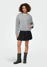 Veste bomber zippée grise, jupe plissée noire et bottes noire hauteur genou avec des accents de boucle. Le matériau semble doux, avec une coupe décontractée.