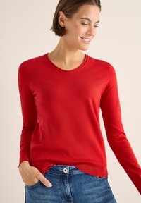 Roter Pullover mit langen Ärmeln und rundem Halsausschnitt, weicher Textur und tailliertem Design, kombiniert mit Jeans aus Denim, die mit einem Knopfdetail versehen sind.