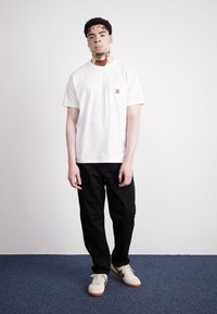 Vit bomull t-shirt med bröstficka och logotyp, tillsammans med svarta byxor och ljusa sneakers på en blå mattbakgrund.