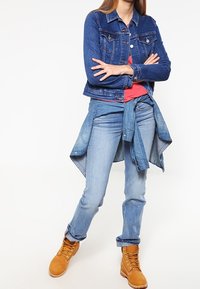 Džínová bunda v tmavě modré barvě nošená přes červenou košili, doplněná o světle modré džíny a béžové boty. Bunda má klasické kapsy a knoflíky.