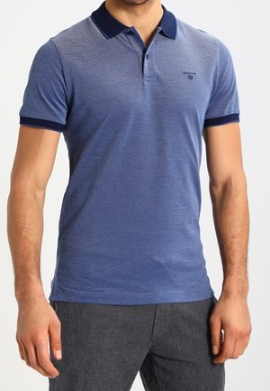 Homme portant un polo bleu à manches courtes avec col et poignets bleu marine, associé à un pantalon gris, debout devant un fond blanc.