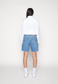 Witte blouse met lange mouwen en blauwe spijkershorts met rafelige zoom. Shorts hebben twee achterzakken en lichte slijtage. Witte sneakers en sokken zichtbaar.