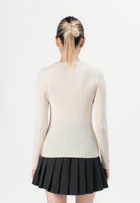 Kvinne med blond hestehale iført en beige teksturert langermet topp og et svart plisserte skjørt, stående mot en ensfarget hvit bakgrunn.