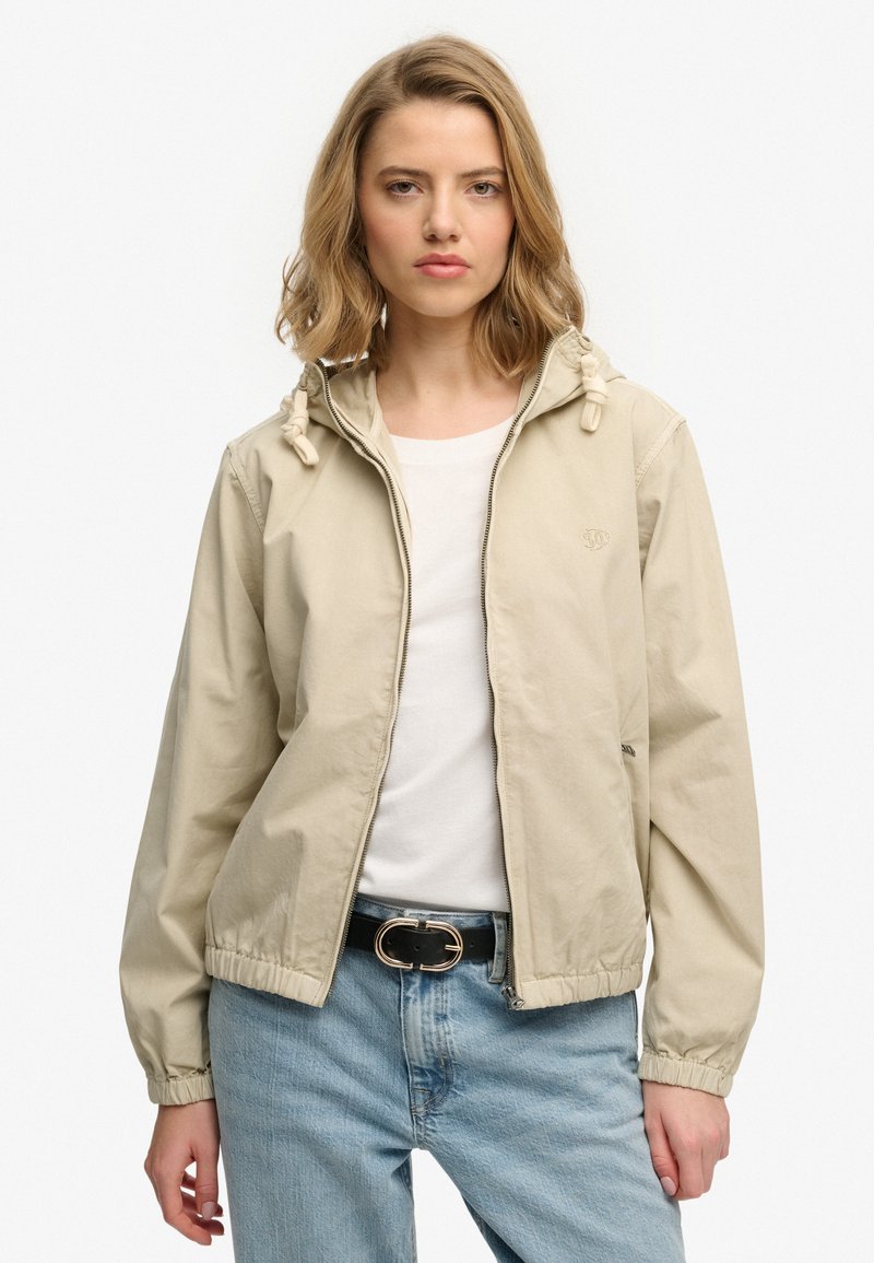 Chaqueta beige claro con cremallera y capucha, puños elásticos y cordones, combinada con una camiseta blanca y jeans azules para un look casual.