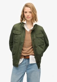 Olijfgroene jas met rits en drukknoopsluiting, vier voorkruiden en een kraag. Draagt over een bruine trui en een wit shirt.