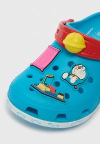 Plave Crocs sandale s crvenim remenom, šarenim dekorativnim elementima, rupama za ventilaciju i bijelim uzorkom oblaka oko potplata.