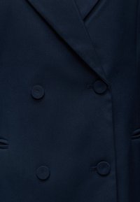 Gros plan d'un blazer bleu marine foncé avec des boutons croisés et de larges revers sur un tissu texturé.