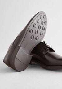 Zapatos de cuero marrón con un acabado brillante, que presentan una suela de goma texturizada con patrones circulares y un tacón bajo. Diseño con cordones y detalles de costura.
