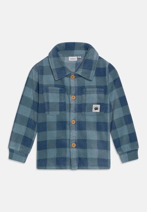 Chaqueta de forro polar a cuadros en azul y verde azulado con botones de madera, dos bolsillos en el pecho y un pequeño parche con una huella de pata en el bolsillo derecho.