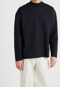 Schwarzes langärmeliges T-Shirt aus Baumwolle mit hohem Kragen, das ein dezentes Logo auf der Brust und eine lockere Passform aufweist.