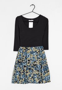 Svart långärmad topp med rund halsringning, kopplad till en blommig plisserad kjol med blå och gröna blommor på en svart bakgrund.