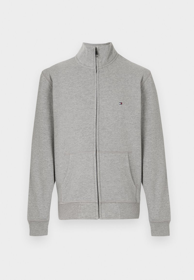 Sudadera gris con cremallera y cuello alto, dos bolsillos frontales, puños de canalé y un pequeño logo en la parte superior izquierda. Textura suave.