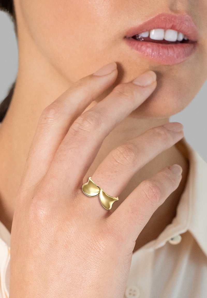 Nahaufnahme einer Hand mit einem goldenen, doppelt blattförmigen Ring am Mittelfinger nahe leicht geöffneten Lippen und einem beigen Hemdkragen mit Knöpfen.