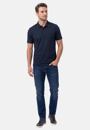 Mies seisoo yllään tummansininen lyhythihainen pikeepaita, siniset farkut ja tummat lenkkarit, toinen käsi taskussa.