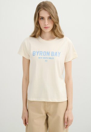 Mujer joven con cabello claro que lleva una camiseta crema con "Byron Bay New South Wales AUS" en azul, combinada con pantalones beige, sobre un fondo liso.