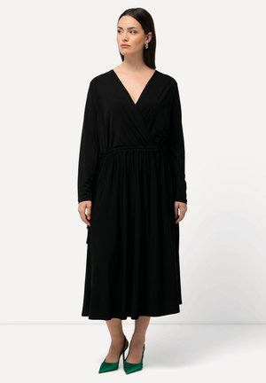 Vestido negro envolvente con mangas largas y cintura ceñida, con un escote en V profundo y falda fluida. Combinado con tacones verdes de punta.