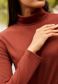 Kvinde, der rører ved sin kraveben, iført en rustfarvet ribbet turtleneck-sweater med lange ærmer udendørs.