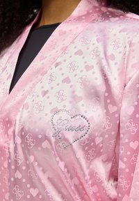 Ρόμπα από ροζ σατέν με μοτίβο καρδιάς και λογότυπο, υφή με πουά και διακοσμητικό από στρας στην αριστερή πλευρά με την επιγραφή "Loves" σε καλλιγραφικό στιλ.