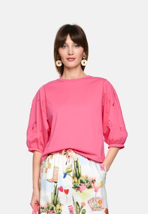 Woman wearing a bright pink blouse with puffed sleeves and white pants featuring colorful desert and cocktail print, posing with one hand in pocket.