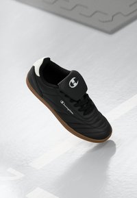 Pantofii sport negri au un superior din piele lucioasă, talpă din cauciuc și un logo alb proeminent pe limbă. Dispune de un guler căptușit și șireturi.