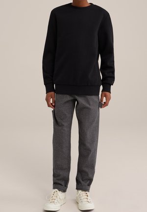 Personne portant un sweat-shirt à manches longues noir, un pantalon gris à carreaux avec des poches latérales, et des baskets blanches montantes, debout sur un fond beige.