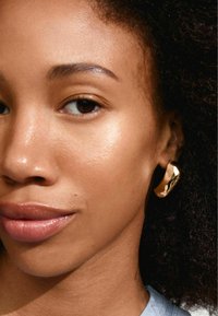 Goldene Creole mit strukturiertem Oberflächenbild, getragen von einer Person. Die Haut zeigt einen natürlichen Glanz, der die reflektierenden Eigenschaften der Ohringe hervorhebt.