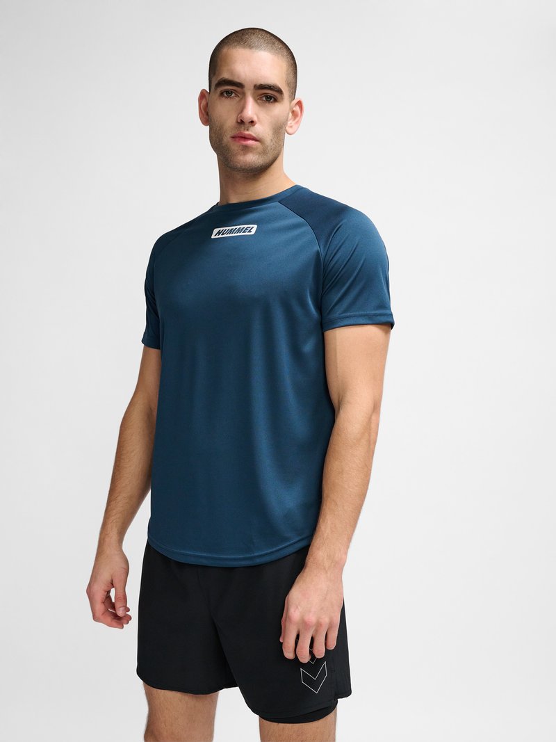 Mand stående fremadrettet iført en blå kortærmet sportstrøje og sorte shorts mod en ensfarvet lys baggrund.