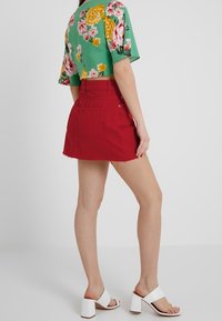 Mini gonna in denim rossa con orlo sfrangiato, abbinata a un crop top verde a stampa floreale. Sandali con tacco a blocco bianchi completano l'outfit.