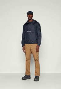 Marineblauer leichter Pullover-Jacke mit einer Fronttasche und Kordelzug, kombiniert mit hellbraunen Hosen und schwarzen Wanderschuhen.