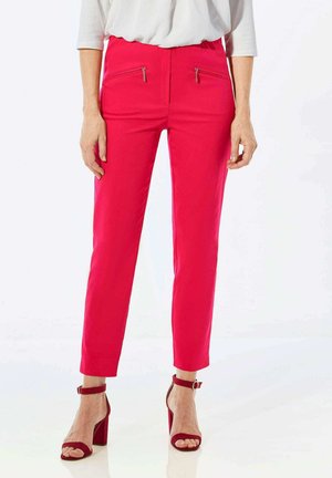 Pantalons roses ajustés avec zips à l'avant ; tissu lisse, design fuselé, longueur raccourcie ; associés à des talons rouges à bride de cheville.