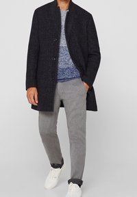 Donker wollen jas met een hoge kraag, knoopsluiting aan de voorkant, gedragen over een blauwe gebreide trui en grijze broek, gecombineerd met witte sneakers.