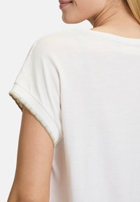 T-shirt en coton blanc avec des manches roulées et une bordure texturée vert et crème. Tissu lisse avec une coupe décontractée et un léger éclat.