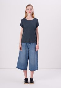 Tums melnā krāsa īsa piedurkne kreklā, apvienota ar platās kājas zilu denim culottes. Apavi ir melni sporta apavi ar brūnas krāsas zoli. Minimālistisks dizains.