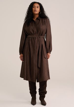 Femme aux longs cheveux bouclés portant une robe marron foncé ceinturée à manches longues et des bottes assorties jusqu'aux genoux, debout devant un fond uni.