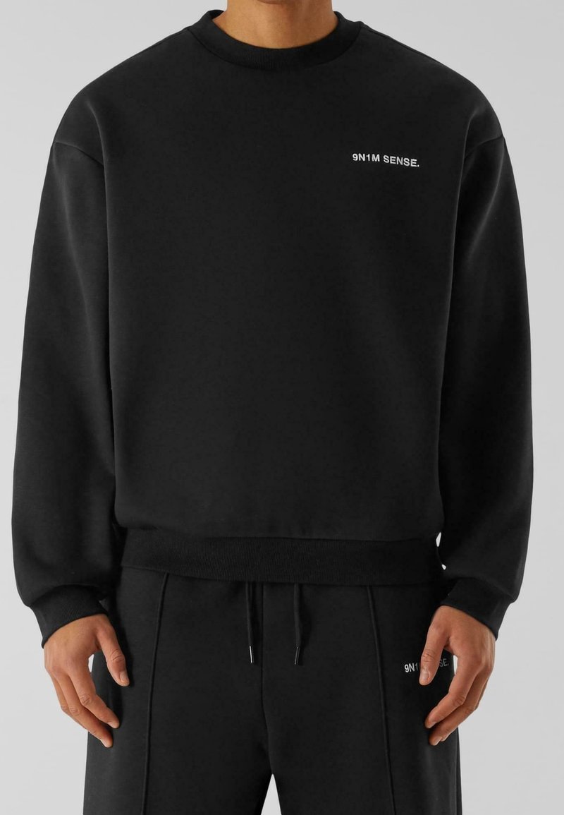 Homme portant un sweat-shirt et un pantalon de jogging noirs, tous deux avec un petit texte blanc « 9N1M SENSE. » sur la poitrine et la cuisse.