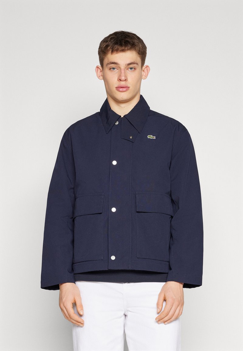 Giacca blu navy con chiusura a bottoni, due tasche anteriori e colletto. Presenta un logo sul lato superiore sinistro.