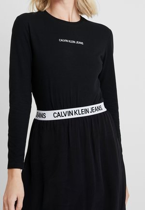 Femme portant une robe noire à manches longues avec un texte blanc "Calvin Klein Jeans" sur la poitrine et une ceinture blanche avec logo assortie à la taille.
