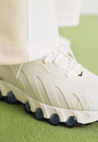 Fehér sportcipő texturált felső résszel, fekete kiegészítő logóval és áttetsző fekete talppal. Zöld háttér előtt elhelyezve.
